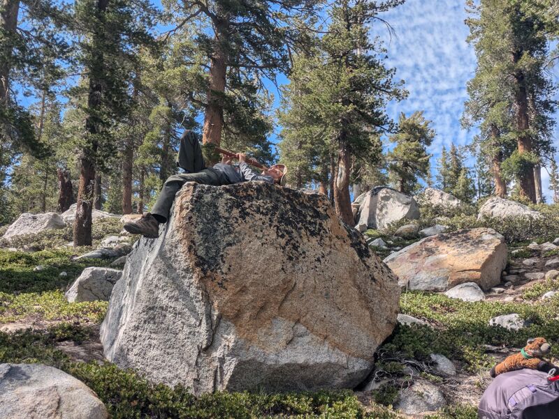 A person is lying on a large boulder in a forest. The person is wearing dark pants, a light-colored shirt, and shoes. They are resting with their arms behind their head. The boulder is covered in lichen and moss. The forest is dense with tall trees and a few other rocks are scattered around. The sky is visible through the trees and is blue with some clouds.